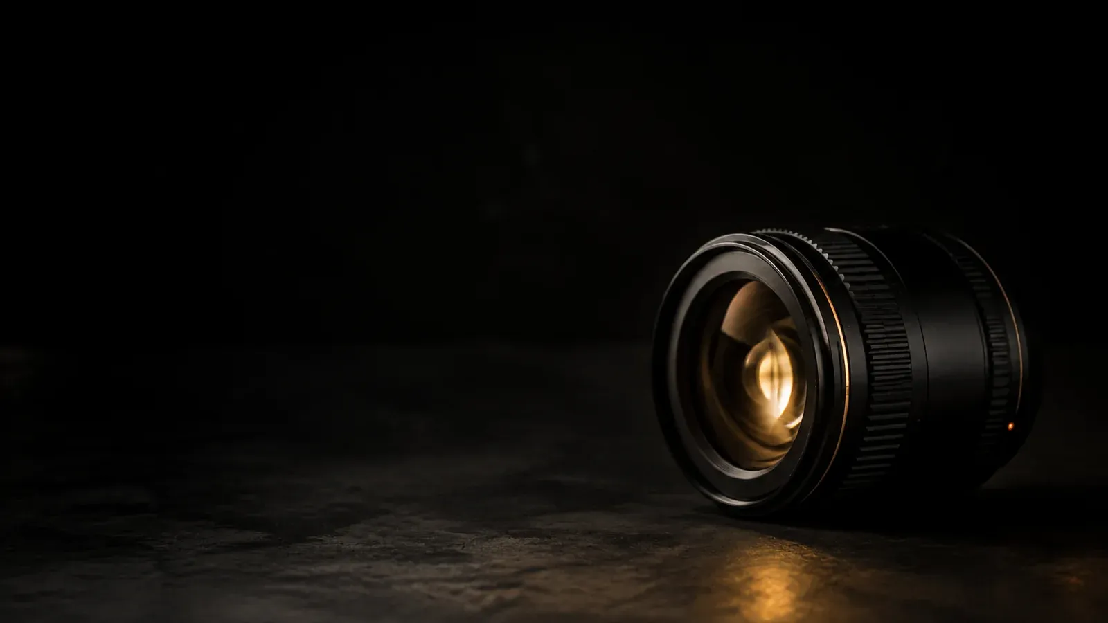 A camera lens standing on a dark surface, a warm light caught in its glass.